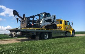 Bock's Service yellow flat-bed hauling truck loaded with a portable winch machine attached to a Bobcat