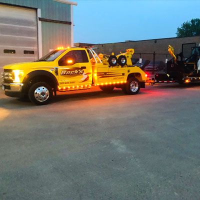 Bock's Service yellow light-duty tow truck towing a trailer with a skidloader on it